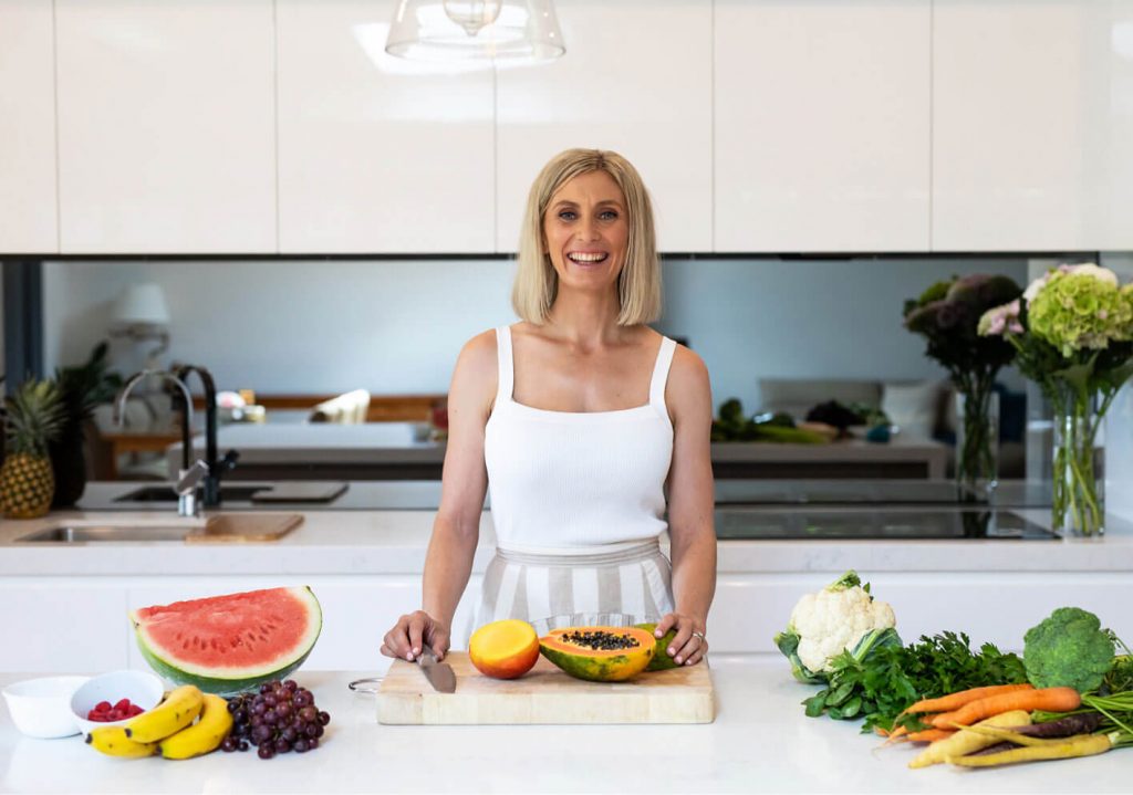 A woman, an accredited practicing dietitian, stands before a cutting board filled with fresh fruits and vegetables.