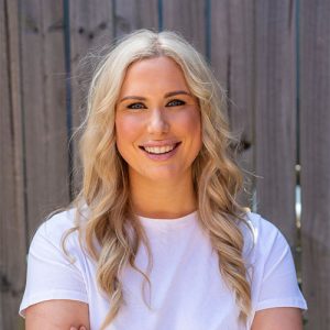 Amanda Smith, a blonde dietitian, poses with arms crossed in front of a wooden fence, symbolizing her focus on women's health.