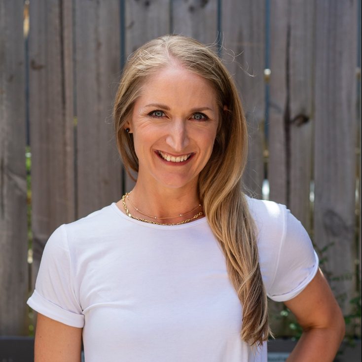 Kate Jones, a clinical dietitian, poses in a white t-shirt and jeans by a wooden fence, embodying a healthy lifestyle.