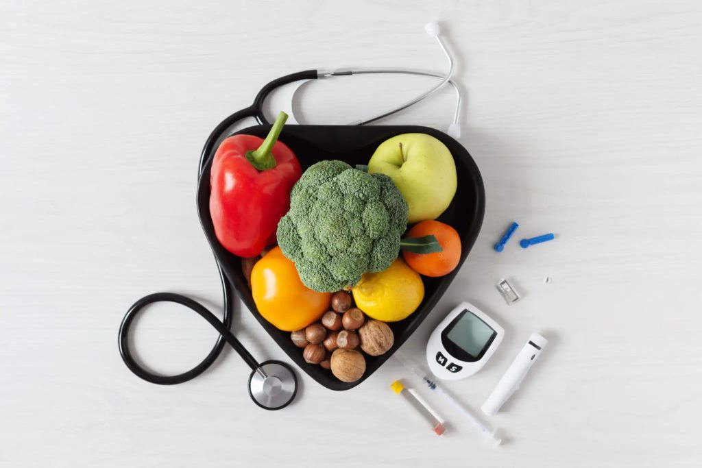 Fresh fruits and vegetables in a heart-shaped bowl with a stethoscope and diabetes monitoring tools, representing chronic disease dietitian support and nutrition plans